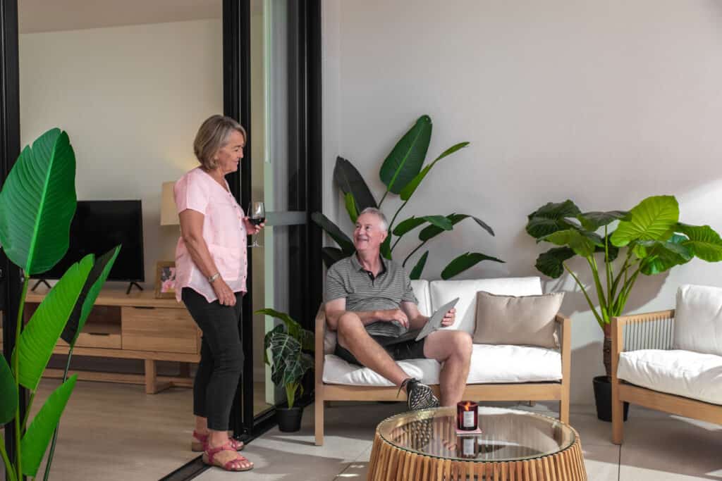 Senior couple are in discussion as the woman stands holding a glass of wine and the man is sitting holding a tablet device.