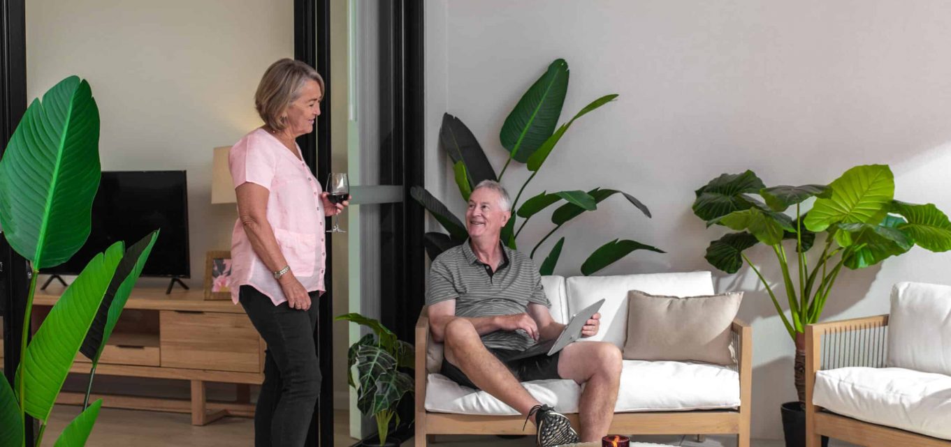 Senior couple are in discussion as the woman stands holding a glass of wine and the man is sitting holding a tablet device.